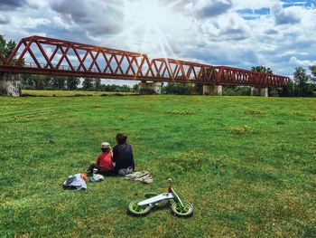 Rear view of mother with son sitting on grassy field against cloudy sky during sunny day