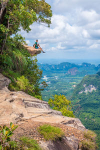 Man sitting in cliff against cloudy sky