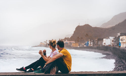 Side view of couple sitting by sea against sky