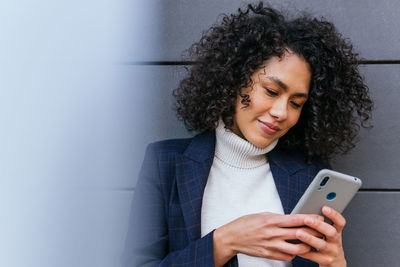 Positive young curly haired ethnic businesswoman reading information on mobile phone while standing near gray wall