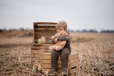 A happy boy with a soft toy is sitting on a box in the field