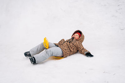 Full body of content kid in outerwear lying on saucer sled while riding down snowy hill on cold winter day