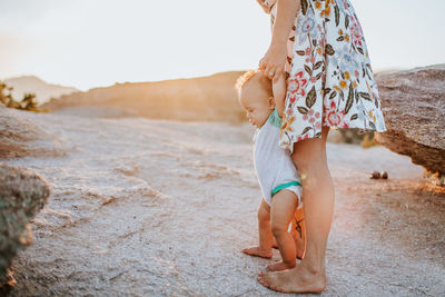 Mom helping baby boy walk on rocks at sunset