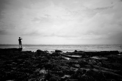 Rear view of man standing on rocks at beach against cloudy sky