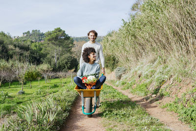 Woman sitting in wheelbarrow, holding fresh vegetables, man pushing her