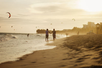 Rear view of couple walking on beach against sky at sunset