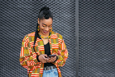 Trendy young ethnic female in colorful clothes browsing on smartphone against gray wall on city street