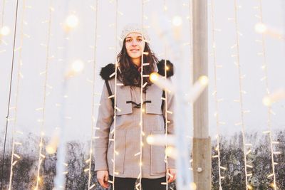 Portrait of woman standing amidst illuminated string lights in winter