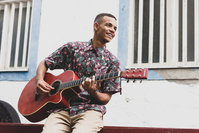 Young man playing guitar while sitting against wall