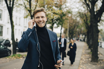 Smiling businessman looking away while holding in-ear headphones in city