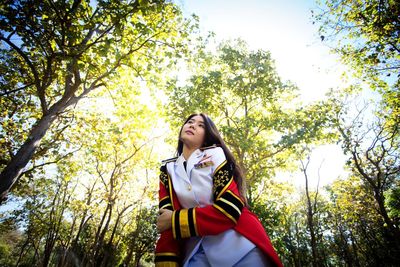 Low angle view of young woman wearing uniform looking away while standing against trees in forest