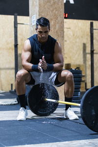 Muscular hispanic male athlete applying talcum powder on hands while sitting near heavy barbell during weightlifting workout in light gym