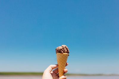 Cropped hand holding ice cream cone against clear blue sky during sunny day