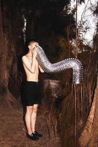 Shirtless man with pipe standing by tree in forest