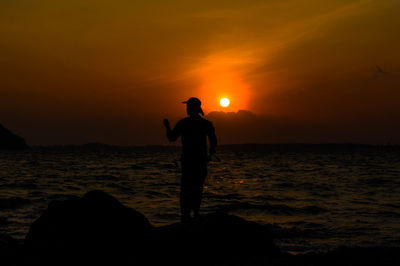 Silhouette man standing on rock by sea against sky during sunset