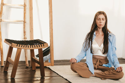 Girl with long and dark hair sitting lotus pose meditating on floor light room interior. 