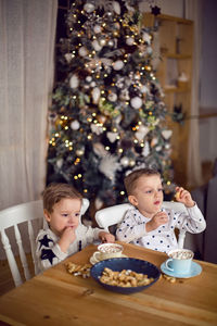 Two brothers in pajamas sitting at  table and drink cocoa with marshmallows and cookies in kitchen