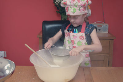 Girl wearing apron straining flour in container on table at home