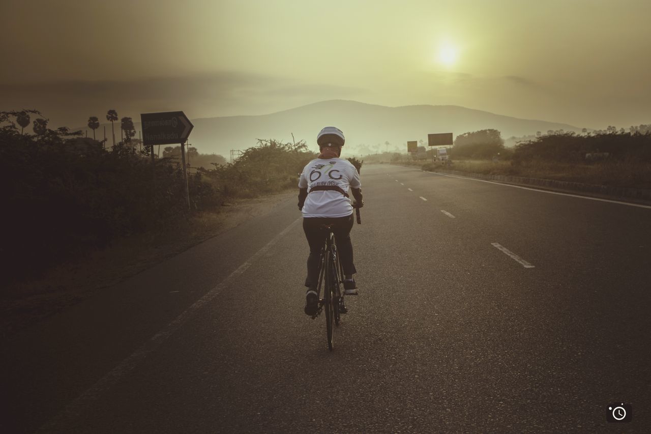 REAR VIEW OF MAN BICYCLING ON ROAD AGAINST SKY