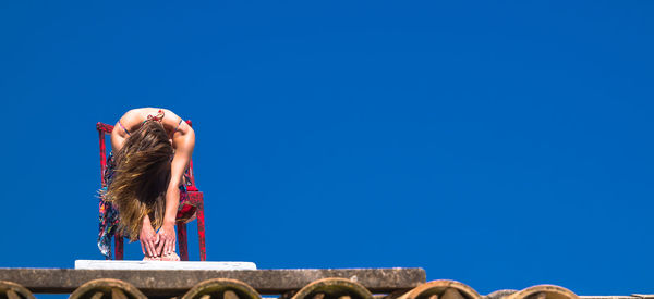 Low angle view of woman sitting on chair at building terrace against clear blue sky