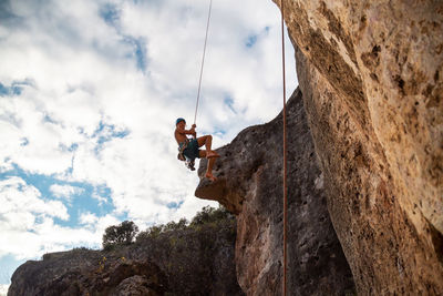 Low angle view of man hanging from safety harness by rock formation against sky