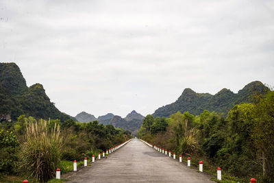 Empty road along trees and mountains against sky