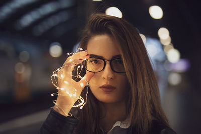 Close-up portrait of young woman holding illuminated string lights while standing at railroad station platform at night