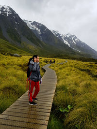 Woman standing on boardwalk over grassy land against snowcapped mountains