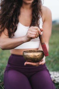 Unrecognizable female athlete in sportswear holding bell and meditating during yoga session in nature