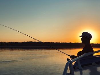 Silhouette man fishing in water against orange sky