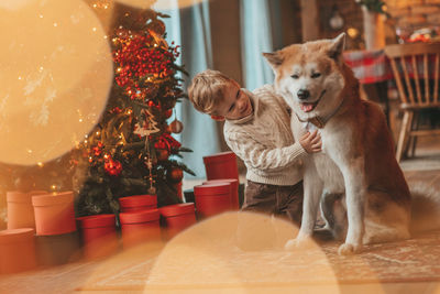 Candid authentic happy little boy in knitted beige sweater hugs dog with bow tie at home on xmas