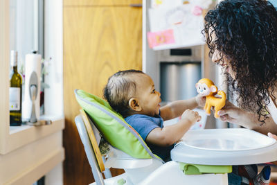 Mother playing with son sitting on high chair in kitchen at home