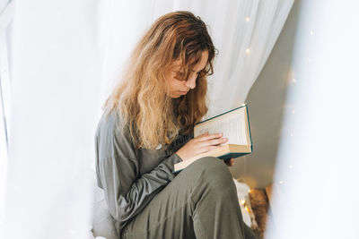 Beautiful teenager girl reading book sitting on windowsill in the cozy children room