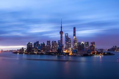 Illuminated city by sea against sky at sunset