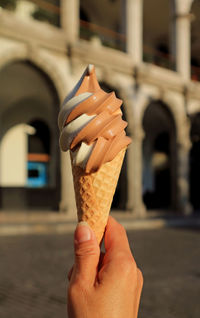 Woman's hand holding a two-tone soft serve ice cream cone, with vintage building in background