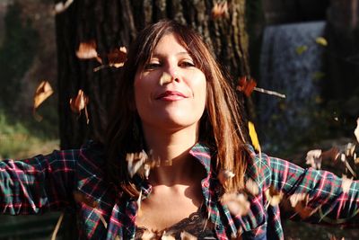 Portrait of woman throwing leaves while standing against tree