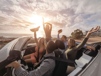 Friends with arms raised sitting in convertible against sky during sunset