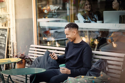 Cheerful young man using smart phone sitting with coffee on bench against cafe window