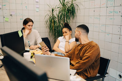 Young businessman sharing smart phone with female colleagues while sitting at creative office