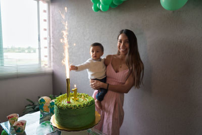 Delighted ethnic mom holding baby boy while celebrating birthday with festive cake decorated with burning firework