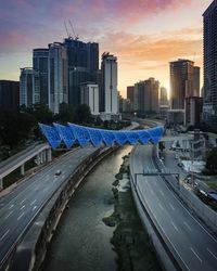 Road by buildings in city against sky during sunset