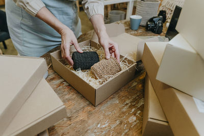 Hands of mature craftswoman packing crochet decor in box on workbench