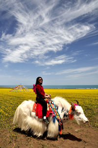 Side view of young woman riding yak on field against sky