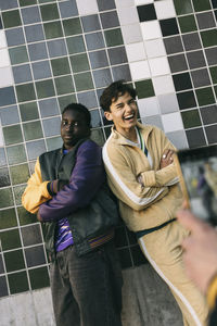 Young man laughing while standing with male friend against tiled wall