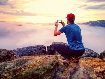 Hiker takes selfie photo, fall nature adventure. man sit on mountain sumit daybreak above milky fog