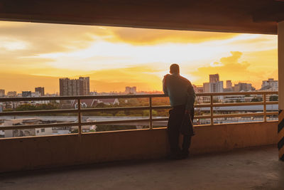 Rear view of man standing by railing against sunset sky