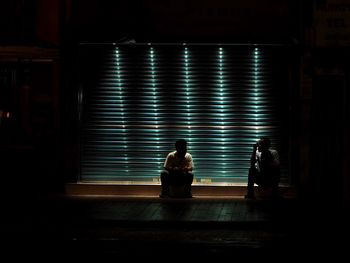 Men sitting against illuminated shutter in city at night