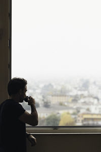 Side view of man drinking by window against cityscape during rain