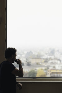 Boy standing by window against clear sky