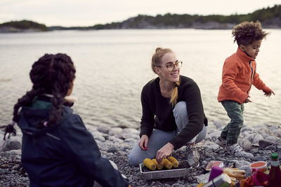 Smiling mother kneeling while cooking corns on barbecue grill amidst children at beach